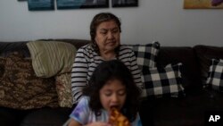 Marina Maalouf, a longtime resident of Hillside Villa, sits on a sofa as her granddaughter eats pizza for lunch in their apartment in Los Angeles, Oct. 1, 2024.